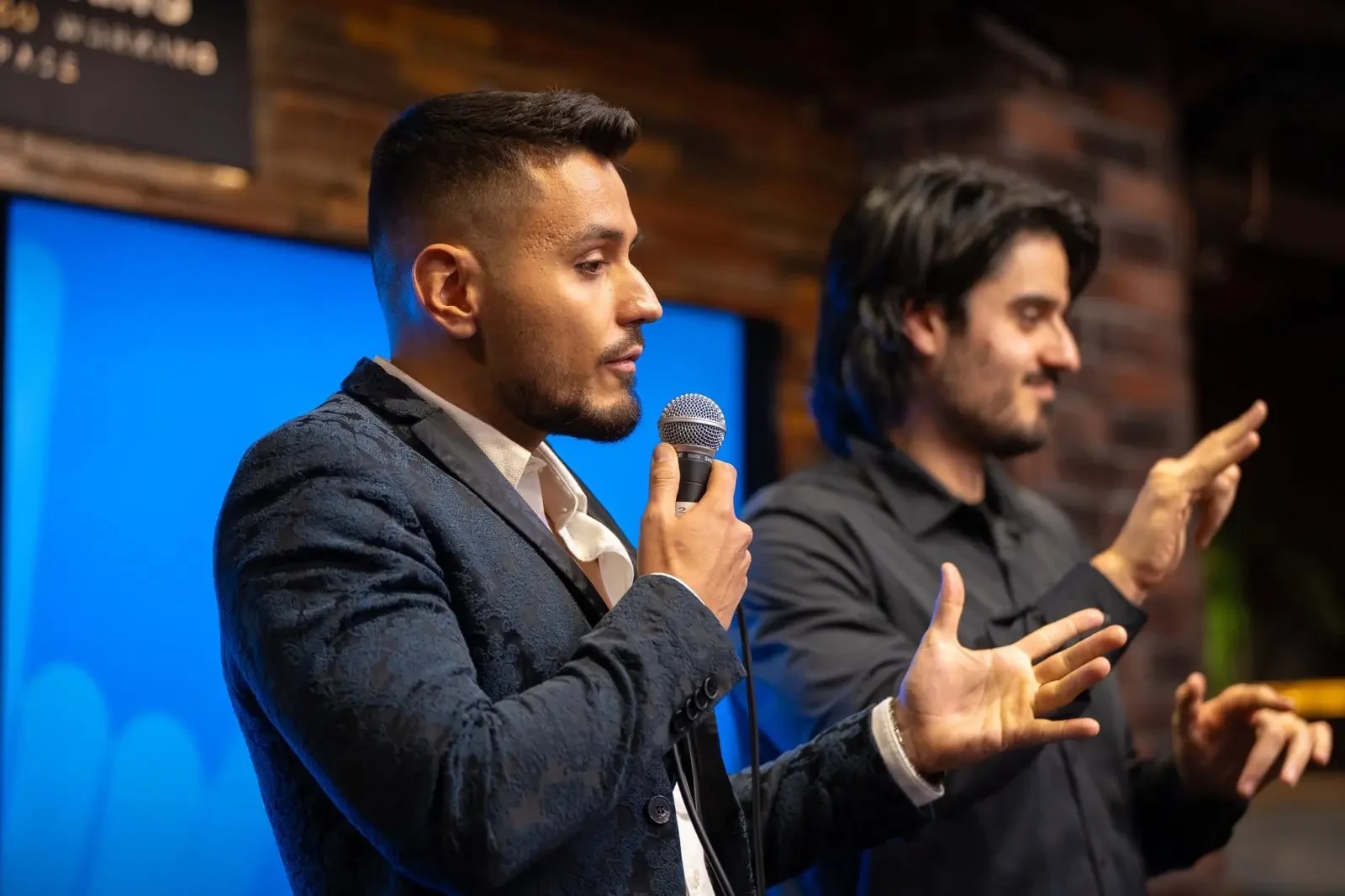 A speaker with a microphone; another person gestures using sign language in the background.
