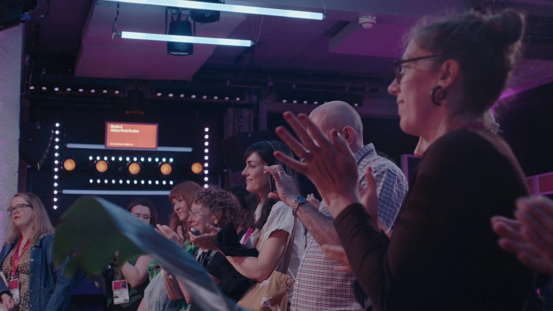 Audience applauding in a studio with colorful lights and stage backdrop.