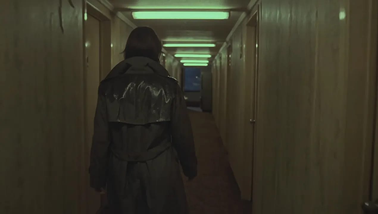 Slow handheld tracking shot behind a detective walking down a narrow motel hallway at midnight, her wet trench coat dripping a faint trail on the carpet. She passes three numbered doors, each closer than the last. As she passes the second door, the fluorescent tube above her flickers and dies, dropping her into shadow for a beat before the next tube buzzes on. She slows, fingertips grazing the peeling wallpaper, and glances at a room-service tray abandoned on the floor, a knocked-over glass still rocking slightly. Near the end of the hall, the vending machine rattles louder and its light stutters as she passes it. In the final seconds, the last door creaks open, spilling a slice of blue TV light across the floor. She freezes, one hand slipping into her coat pocket. The shot ends holding on her still silhouette against the light. Audio: muffled footsteps on damp carpet, a fluorescent buzz that cuts out then returns, the vending machine's uneven rattle growing closer, distant rain, one sharp door creak, a TV murmuring behind a wall, no music. 1970s neo-noir, 35mm film grain, warm green fluorescent cast mixed with cold blue spill from the open door, realistic motion. No readable text, no logos, no extra people on screen.